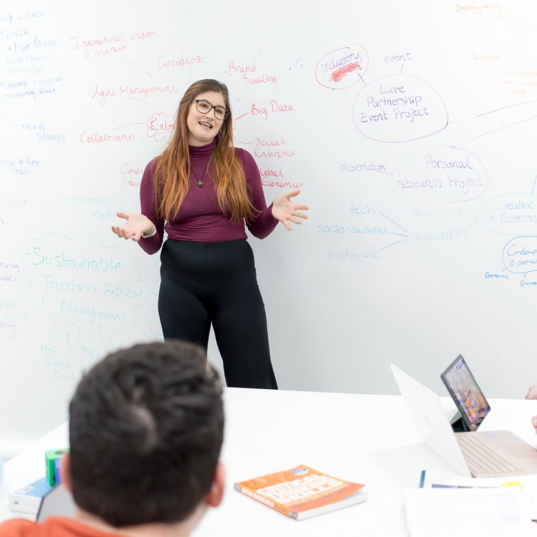 A student leads a discussion, with a large whiteboard full of notes behind her.